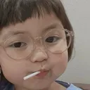 A young child wearing glasses looks sideways with a judgmental expression while holding a lollipop in their mouth.