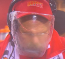 Andy Reid wearing a red hat and a clear plastic face shield looking intensely forward.