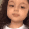 A young child with curly hair looks at the camera with a serious expression while giving a thumbs up.