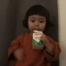 A young toddler with a bob haircut stares blankly while drinking from a small carton of milk.