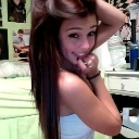A young woman with long brown hair smiling coyly while touching her face with her hand.
