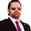 A man wearing a black suit, white shirt, red tie, and dark sunglasses looking directly at the camera with a serious expressi…