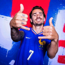 Antoine Griezmann smiling and making the shaka hand gesture while wearing a blue French national team jersey.