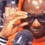 Man wearing an orange hoodie squinting while adjusting his glasses in front of a microphone.