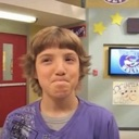 A young boy with brown hair wearing a purple shirt makes a skeptical face with pursed lips.