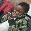 A young boy in a camouflage jacket holds a smartphone and looks at the screen with a deeply confused and skeptical express...