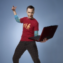 Sheldon Cooper stands holding a laptop while gesturing with his hand in a confident manner.