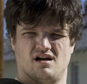 Man with curly brown hair squinting his eyes and looking skeptical with his mouth slightly open.