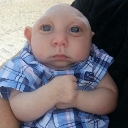 Baby with wide eyes making a heart shape with its hands while wearing a blue plaid shirt.