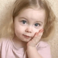 A toddler rests their face in their hands with a shy and bashful expression.
