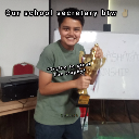 A young boy smiling widely while holding a gold trophy in a classroom setting.