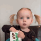 A toddler with pigtails eating a snack from a box while looking around.