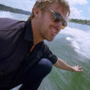 Ryan Gosling wearing sunglasses and a black shirt, reaching his hand out over the water with a relaxed expression.