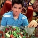 A young boy in a blue shirt stares at the camera with a skeptical expression while holding a bowl of salad.