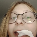 A person wearing glasses looks directly at the camera with a piece of white paper held between their teeth.