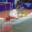 Human head with light blue hair resting on a table next to fruit and a small red figurine.