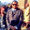 Man in a suit and sunglasses standing with a smug, indifferent expression.