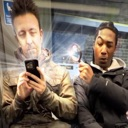 Two men on a train with one using a magnifying glass to look at the other's phone screen.