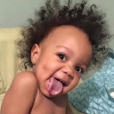 A baby with curly hair sticking their tongue out with a mischievous expression.