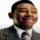 Tyler the Creator wearing a suit and tie with a smug, knowing smile.