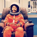 A child wearing an orange astronaut suit and helmet sits in a chair looking prepared for space travel.