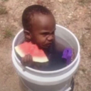 Baby sitting inside a white bucket holding a slice of watermelon with a serious expression.
