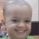 A young child with a shaved head wearing a green shirt, looking directly at the camera with a wide, mischievous, and smug gr…