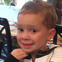 A young boy with brown hair giving a forced, tight-lipped, and pained smile.