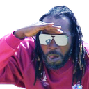 Man with dreadlocks and sunglasses wearing a red hoodie, holding his hand to his forehead to squint into the distance.