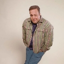 Kevin James wearing a plaid shirt and blue jeans leaning forward with a smug, knowing smirk.