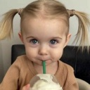 A toddler with pigtails sips from a cup with a straw while looking directly at the camera with a smug expression.