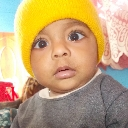 Baby wearing a yellow beanie looking directly at the camera with a confused expression.