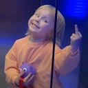 A young child in an orange shirt smiles cheekily while holding up their middle finger.