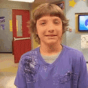 A young boy with shaggy hair giving a hesitant, awkward smile in a classroom setting.