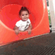A toddler in a white shirt waves goodbye while sliding into a bright orange playground tunnel.