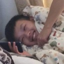 A young boy lying in bed under a blanket, laughing while looking at a smartphone.
