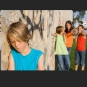 A sad boy stands alone in the foreground while a group of children laugh and point at him in the background.