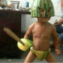 A small child wearing a watermelon rind helmet and loincloth stands in a fighting stance while holding a wooden sword.