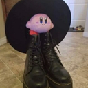 Kirby plush toy wearing a large black hat and sitting on black combat boots.