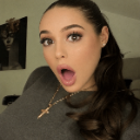 A woman with a shocked expression, mouth wide open and eyes widened, wearing a gold cross necklace.