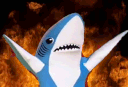 A blue plush shark with arms raised in front of a background of intense fire.
