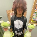 A person with shaggy brown hair holding two green frog plushies.
