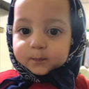 A toddler with wide, curious eyes wearing a dark patterned headscarf looks directly at the camera.