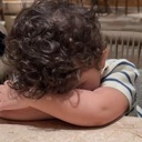 A toddler with curly hair rests their head face-down on their crossed arms in a sign of exhaustion.