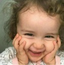 A toddler with curly hair smiling brightly while holding their face with both hands.