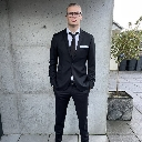 Man in a black suit and tie standing against a gray concrete wall with a neutral expression.
