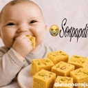 A baby smiling while eating a yellow, cube-shaped Indian sweet.