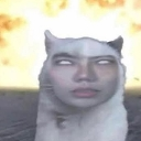 A cat with a human face and blank white eyes stands in front of a massive explosion.