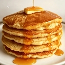 Pancakes stacked high on a white plate with a pat of butter and syrup drizzling down the sides.