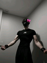 A person wearing a black mask with a bright pink bow standing in a dimly lit room.
